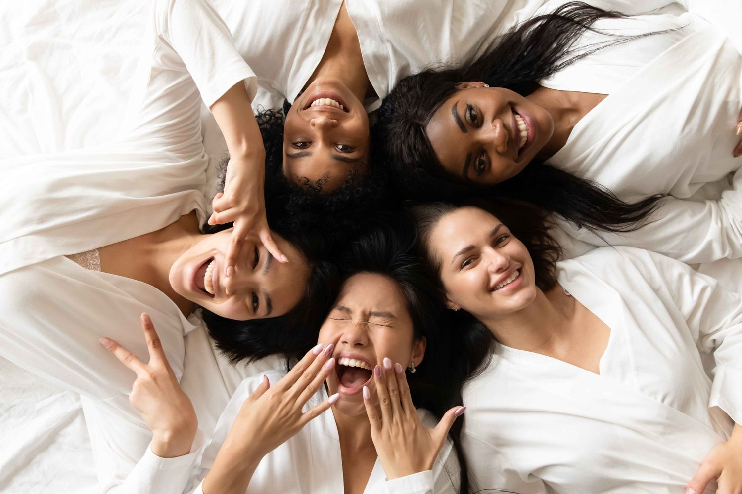 Five friends enjoying laughter on a white bed.